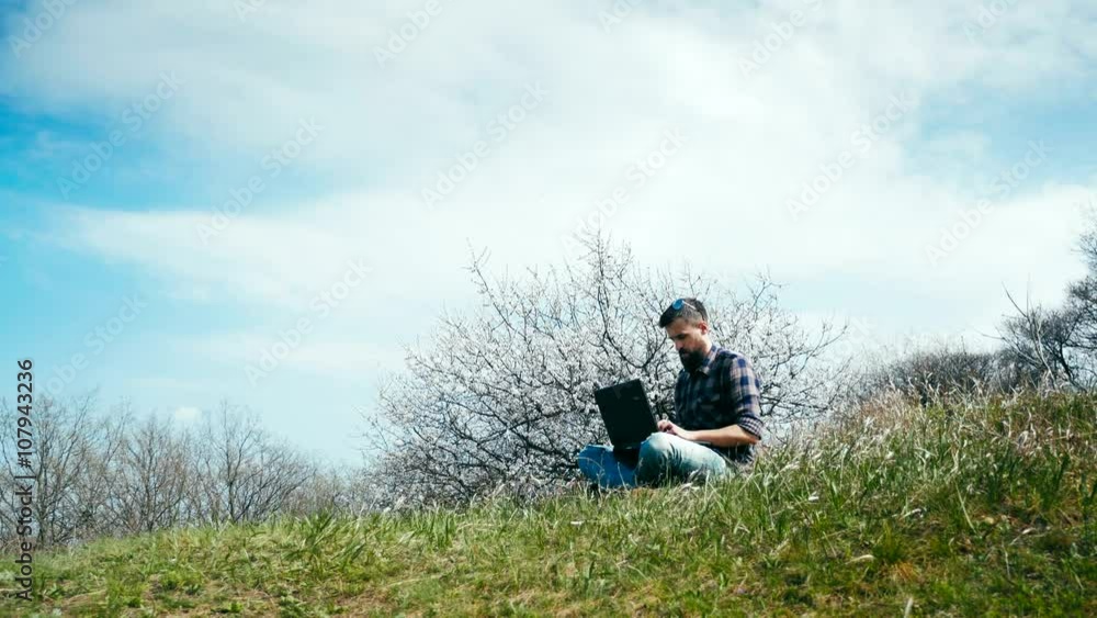 Dolly shot. Man with a beard uses laptop on nature near blossoming tree Dolly shot. Man with a beard uses laptop on nature near blossoming tree