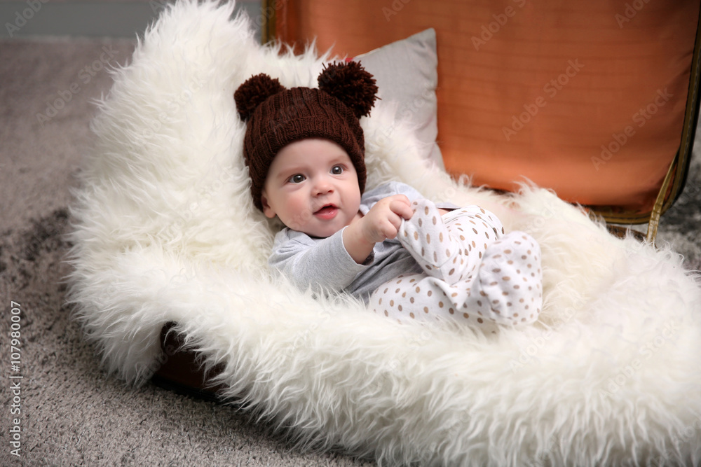 Fototapeta premium Adorable baby in brown knitted hat in luggage bag in the room, close up