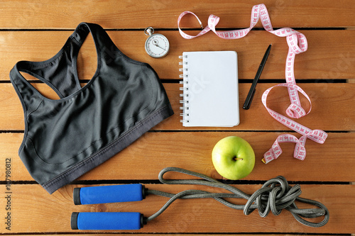 Athlete's set with female clothing, equipment, apple and notebook on wooden background