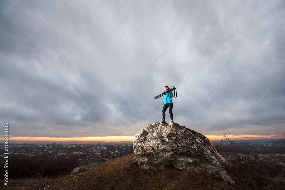 Young attractive female photographer is standing with her camera on tripod on the big rock at city overview point at the sunset. Girl is holding tripod on her shoulder. Blue clouds in the background