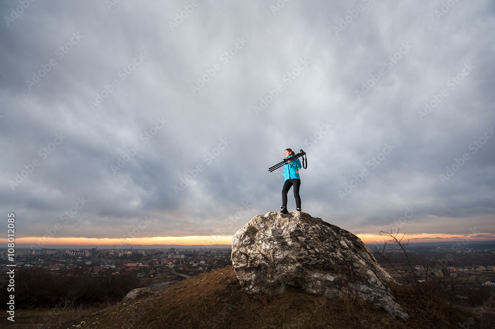 Young attractive female photographer is standing with her camera on tripod on the big rock at city overview point at the sunset. Girl is holding tripod on her shoulder. Blue clouds in the background Young attractive female photographer is standing with her camera on tripod on the big rock at city overview point at the sunset. Girl is holding tripod on her shoulder. Blue clouds in the background