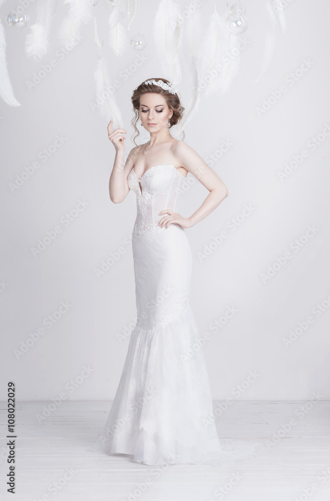 Exquisite young and dreamy bride in a long luxurious lace wedding dress. She is elegant and delicate. The dress is translucent. White feathers around her. Exquisite young and dreamy bride in a long luxurious lace wedding dress. She is elegant and delicate. The dress is translucent. White feathers around her.