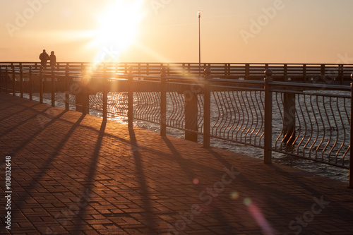 силуэты двух людей, на пирсе, на фоне яркого солнца photo
