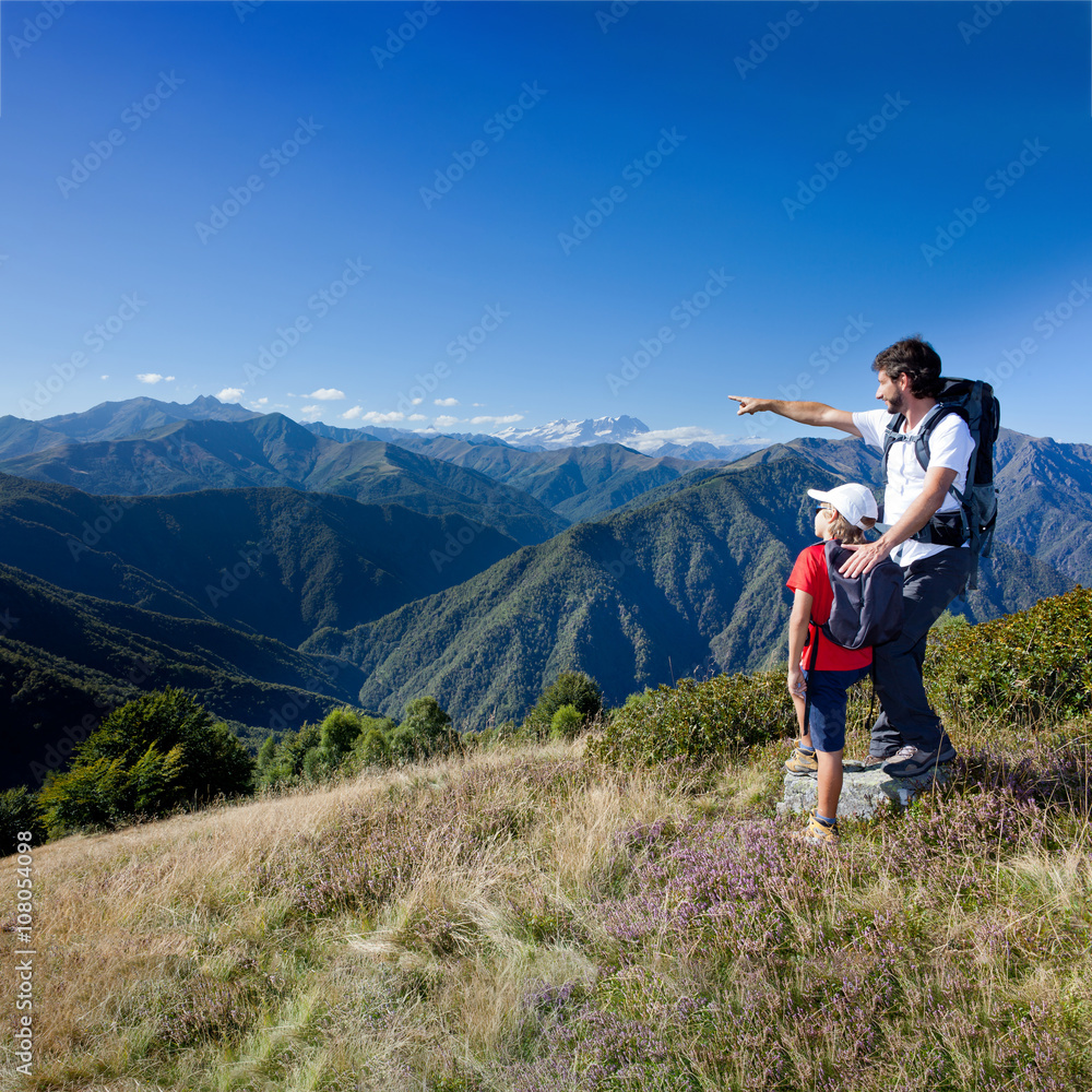 Summer vacation in mountain. Father and young son standing in a mountain meadow. Summer season, clear blue sky. Piemonte, west italian Alps.