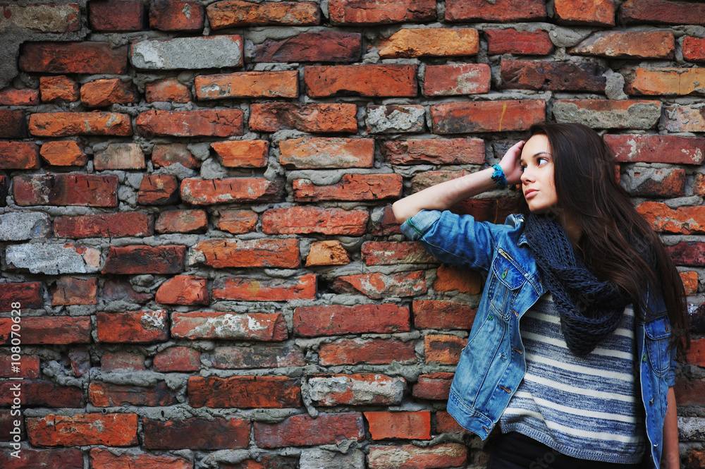 girl with long hair, dressed in a denim jacket and a scarf standing near a wall made of red bricks and waits girl with long hair, dressed in a denim jacket and a scarf standing near a wall made of red bricks and waits