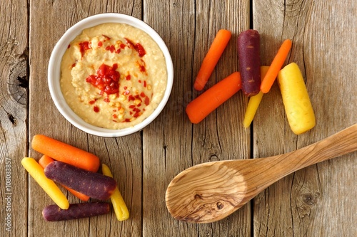 Baby rainbow carrots with hummus dip and spoon, overhead view on a rustic wooden background photo