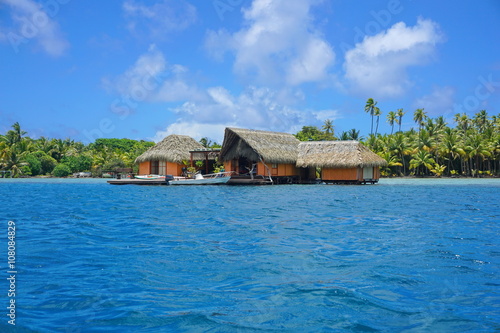 Tropical home with thatch roof over water with the shore of an islet in background, Huahine island, Pacific ocean, French Polynesia