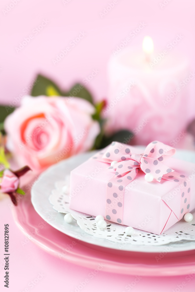 Table place setting with a gift on a plate with roses and candle against pink background