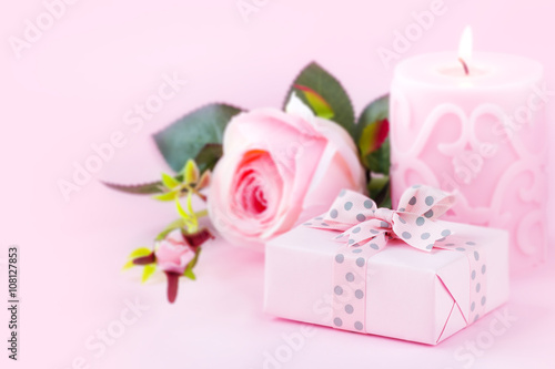 Romantic setting with a wrapped gift, lit candle and a rose flower against pastel pink background. With copy space for your text