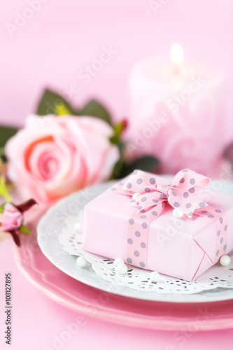 Table place setting with a gift on a plate with roses and candle against pink background