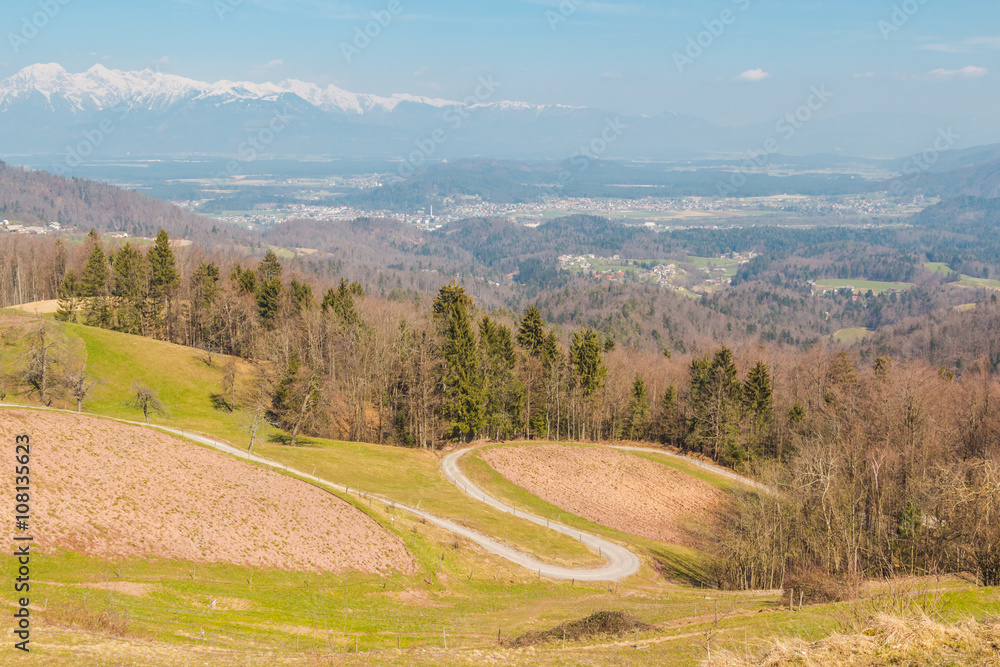 Curving country road on a hillside with a view at a valley and snowy peaks of Kamnik-Savinja Alps.