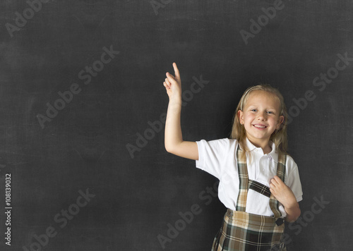  junior schoolgirl with blonde hair standing and smiling happy pointing to copy space on class blackboard