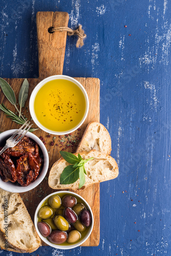 Mediterranean snacks set. Olives, oil, herbs and sliced ciabatta bread on yellow rustic oak board over painted dark blue background