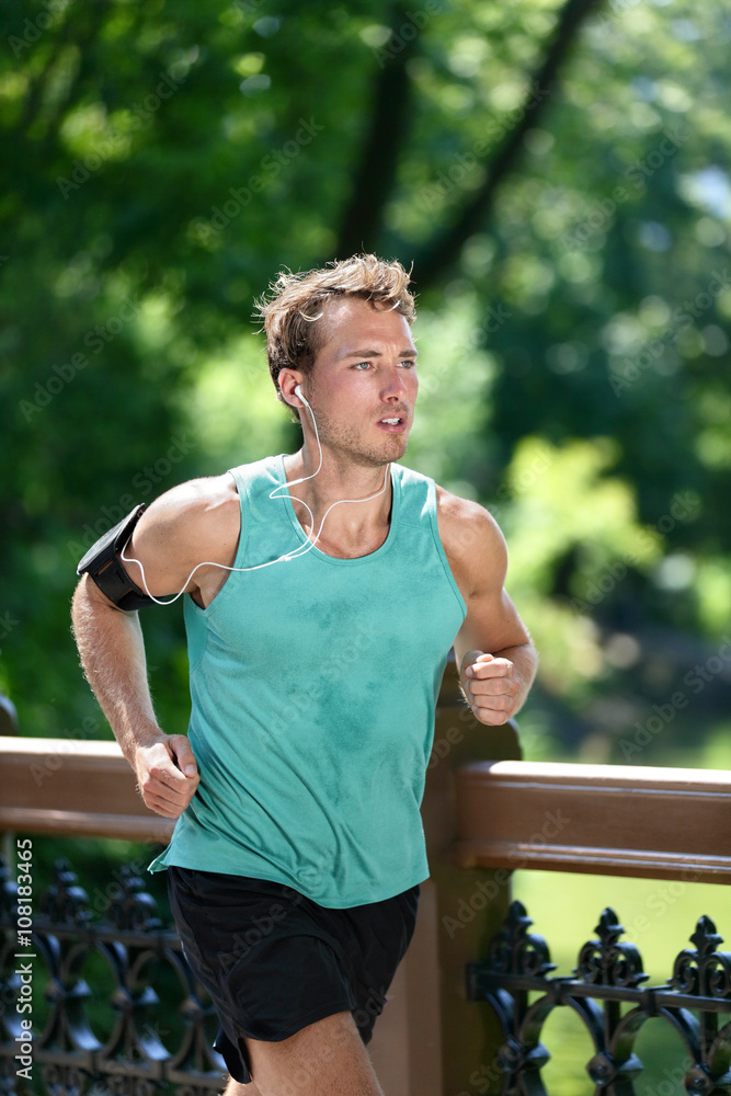 Runner running listening to music with earbuds fitness armband workout app. Male athlete training during summer in urban New York city Central park sweating in sportswear clothing. Photo