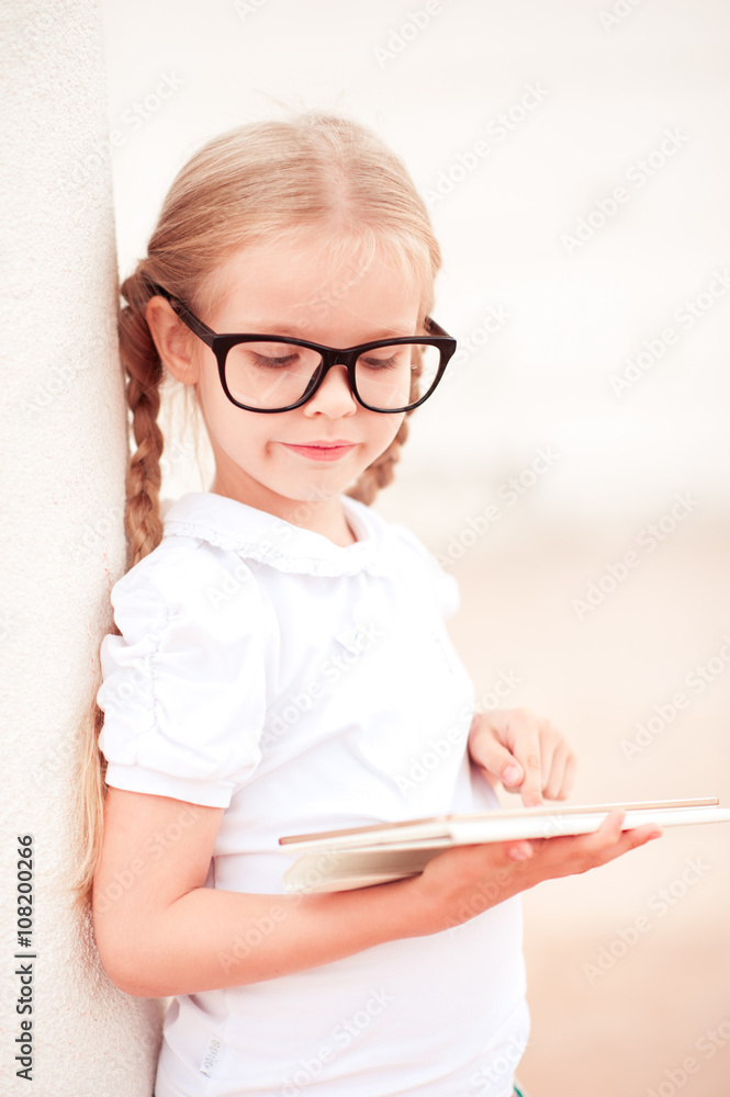 Baby girl 4-5 year old holding tablet computer outdoors. Wearing glasses. Childhood. Baby girl 4-5 year old holding tablet computer outdoors. Wearing glasses. Childhood.