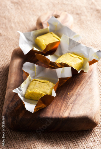 Dehydrated condiment bouillon stock cube salty meat and vegetables aromatic yellow spice, ingredient single whole portion wrapped and open in paper pack , view from above.