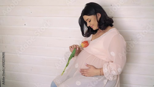 Pregnant beautiful girl smelling a tulip on beige wall background. 4k photo