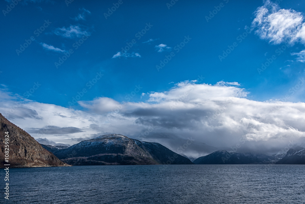 Landscape with ocean and mountain peaks and a blue and cloudy sky.