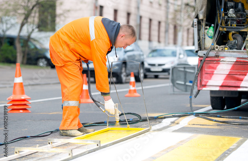 Technical person man worker painting and remarking pedestrian crossing lines on asphalt surface using paint sprayer gun during capital street pavement maintenance works