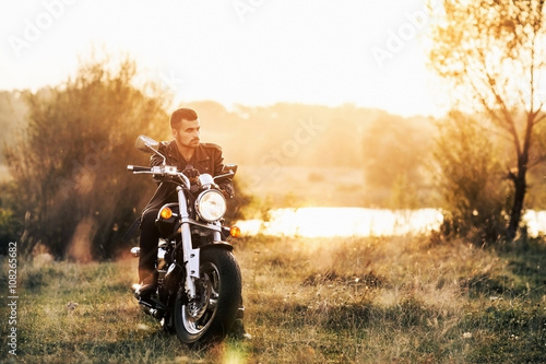 Fotografie young brutal man in a black jacket and glasses sits near a motorcycle