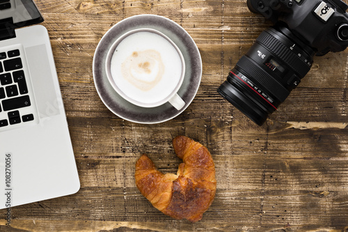 laptop, camera, coffee and croissant on a wooden table