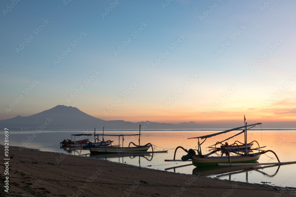 Beautiful sunrise scene at Sanur beach Bali, Indonesia. The rowboat or “Jukung” is widely used as fishing boat by the local fisherman. Mount Agung is distant background. Beautiful sunrise scene at Sanur beach Bali, Indonesia. The rowboat or “Jukung” is widely used as fishing boat by the local fisherman. Mount Agung is distant background.
