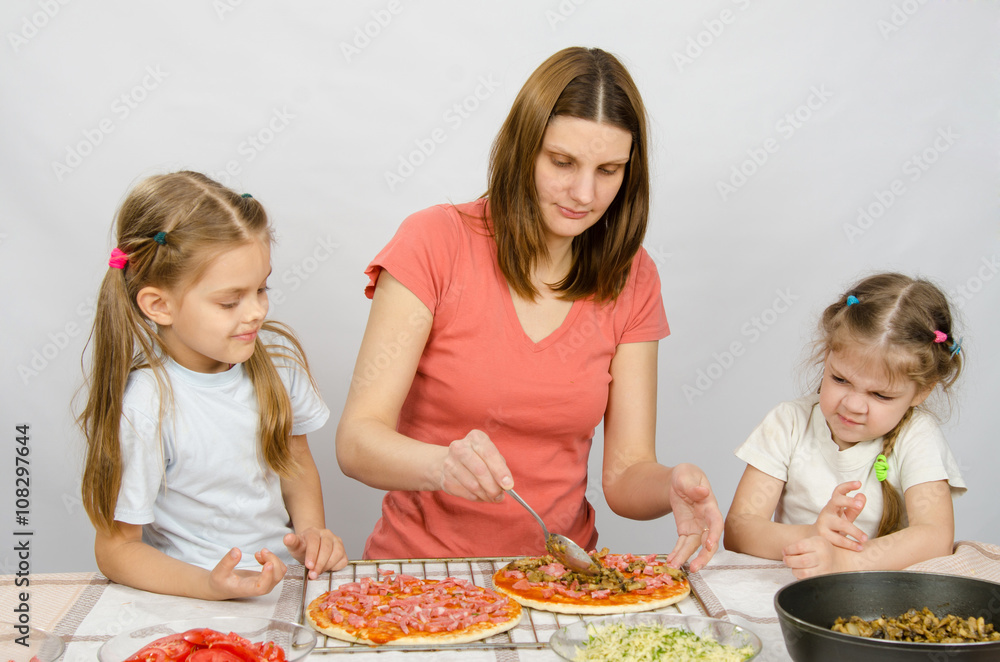 Two little girls sitting at the kitchen table and watch as a mother preparing a pizza