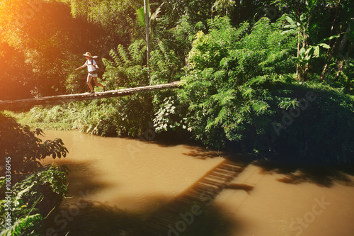 Young woman with backpack walking across hanging bridge above the river in tropical forest  intentional sun glare 