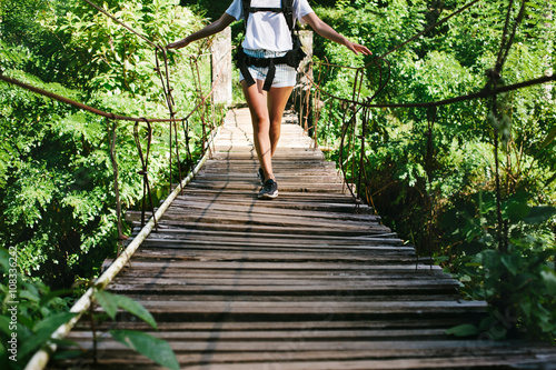 Young fit woman with backpack traveling across hanging bridge in tropical forest