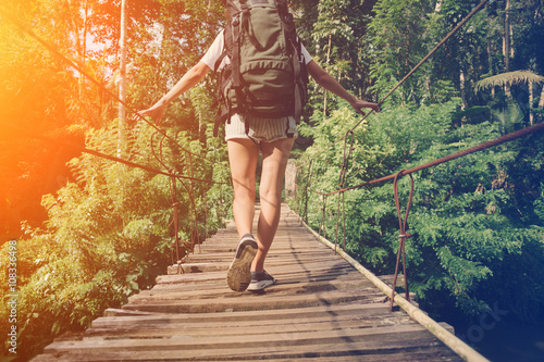 Young woman with backpack balancing across hanging bridge in tropical forest (intentional sun glare)