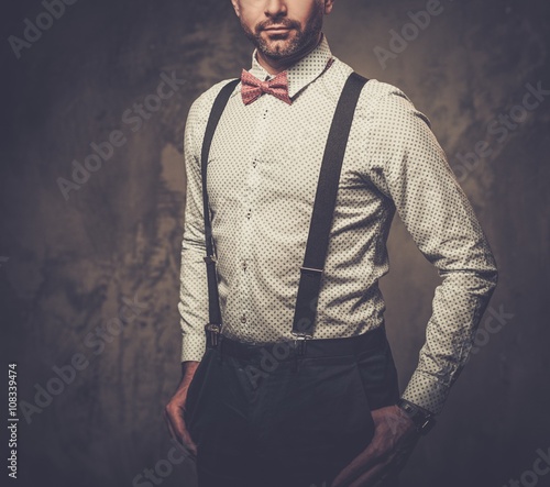 Fotografi Stylish man with bow tie wearing suspenders and posing on dark background
