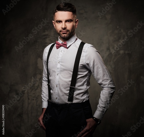Stylish man with bow tie wearing suspenders and posing on dark background.
