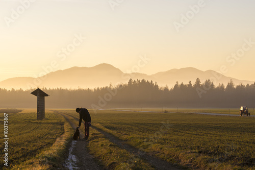 Young man leaning to pet his black dog standing on a country roa