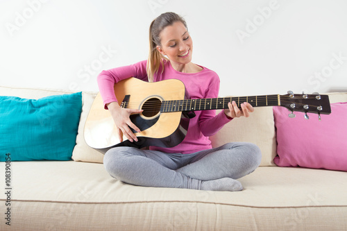 Portrait of a beautiful young woman sitting on a sofa in a living room and playing acoustic guitar