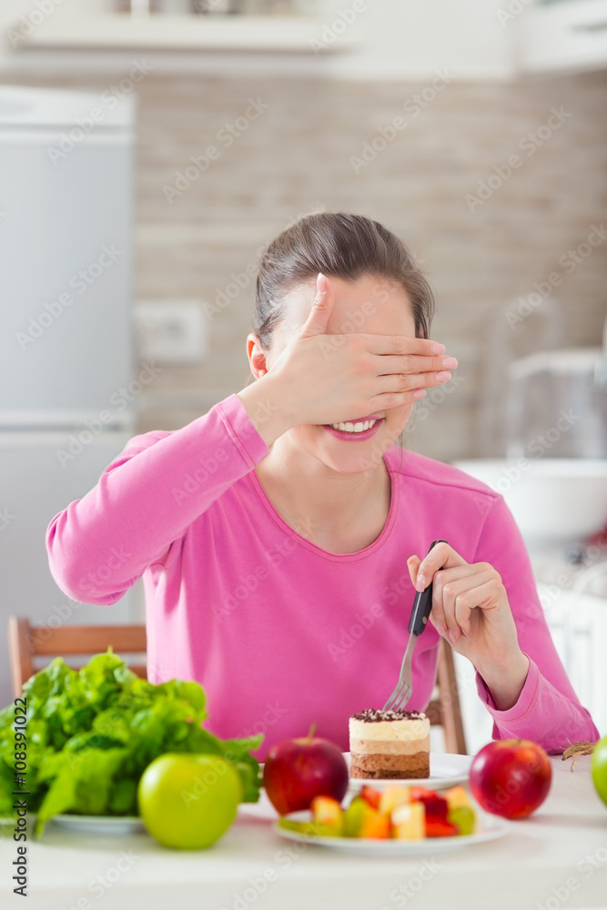 Young woman covering her eyes while eating a cake instead of healthy fresh fruit and vegetable on the table Young woman covering her eyes while eating a cake instead of healthy fresh fruit and vegetable on the table