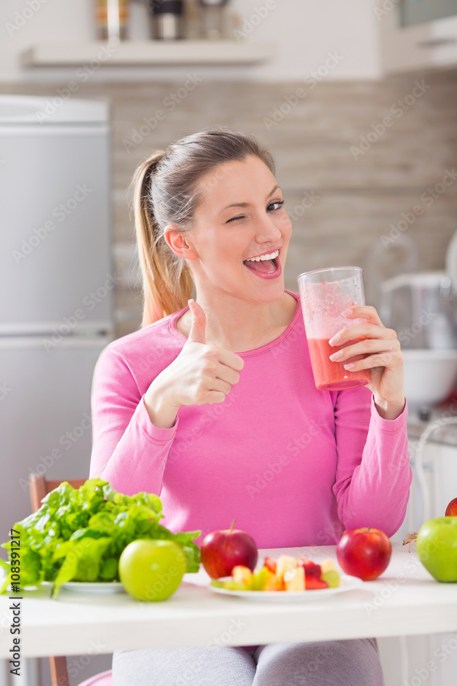 Happy young woman showing OK sign and winking while drinking fresh healthy smoothie in her kitchen