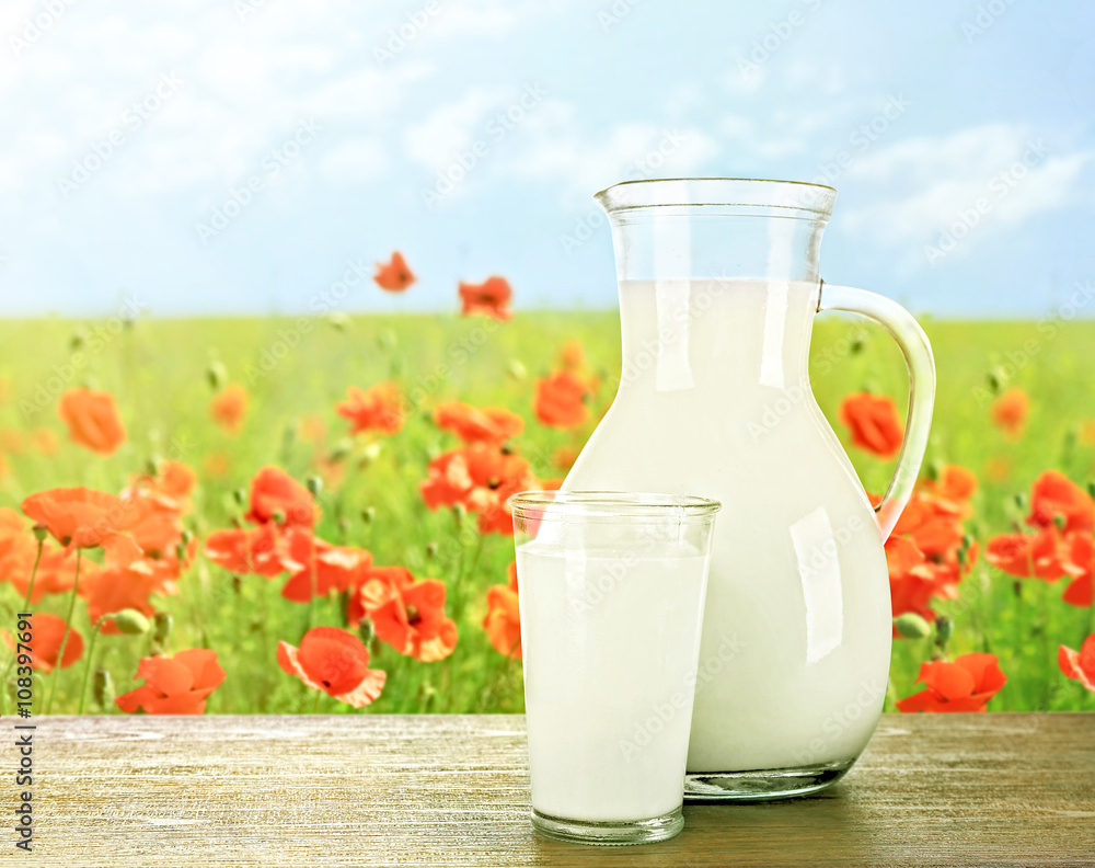 Pitcher, glass of milk on wooden table against poppies and blue sky background Pitcher, glass of milk on wooden table against poppies and blue sky background
