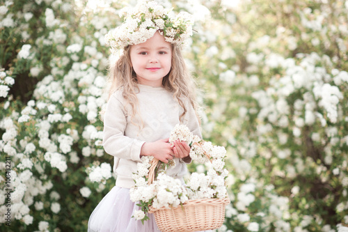 Smiling baby girl holding basket of flowers outdoors. Posing with flower wreath over floral background. Looking at camera. Cheerful. Childhood. Summer time. 