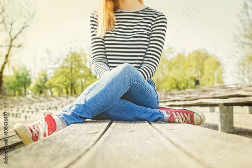 Woman in a blue jeans, striped t-shirt and  red canvas sneakers on her feet, sitting on a bench, legs crossed. Bright and sunny day. © Marina P.