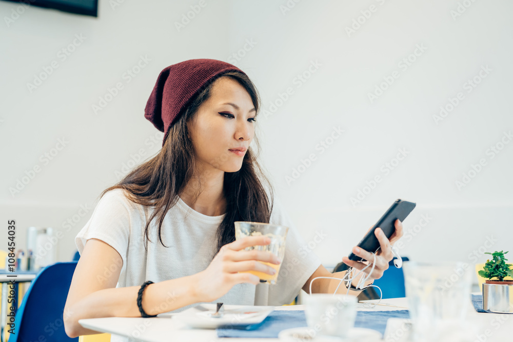 young handsome asiatic  woman sitting in a bar, holding a smart phone and glass of juice, looking down and tapping the screen - technology, social network, communication concept - obrazy, fototapety, plakaty 
