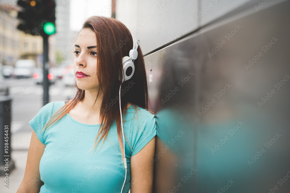 Half length of young handsome caucasian reddish straight hair woman leaning against a wall listening music with headphones, overlooking right, pensive - thinking future, thoughtful concept
