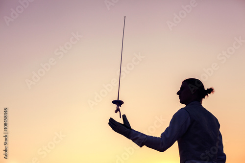 Fencer man wearing white fencing costume and throwing up his fencing sword on a sunset background