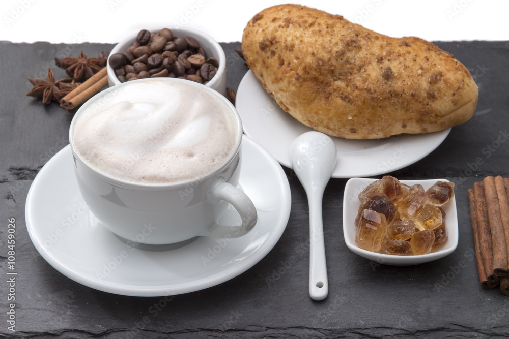 Cup of coffee cappuccino with milk foam on a saucer, fresh baked bun, caramelized sugar, coffee beans, cinnamon. Popular morning Breakfast. on a black background plates of slate Cup of coffee cappuccino with milk foam on a saucer, fresh baked bun, caramelized sugar, coffee beans, cinnamon. Popular morning Breakfast. on a black background plates of slate