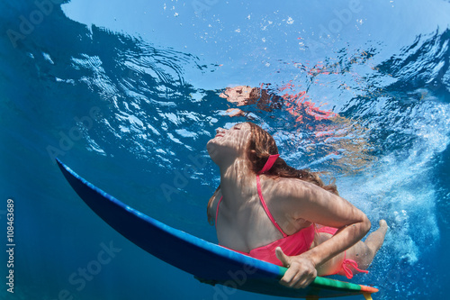 Young girl in bikini in action - surfer with surf board dive underwater with fun under big ocean wave. Family lifestyle, people water sport lessons, beach extreme swimming activity on summer vacation