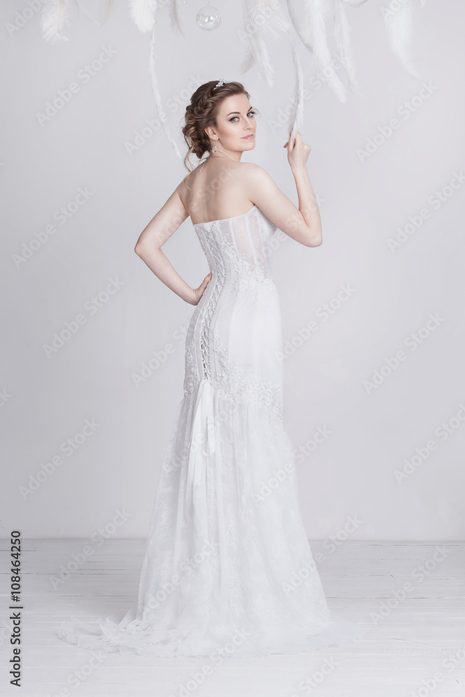 Young and dreamy bride in a luxurious lace wedding dress. She has beautiful dark blond hair and pale skin. She is elegant and delicate. The dress has lacing on the back. White feathers around her.