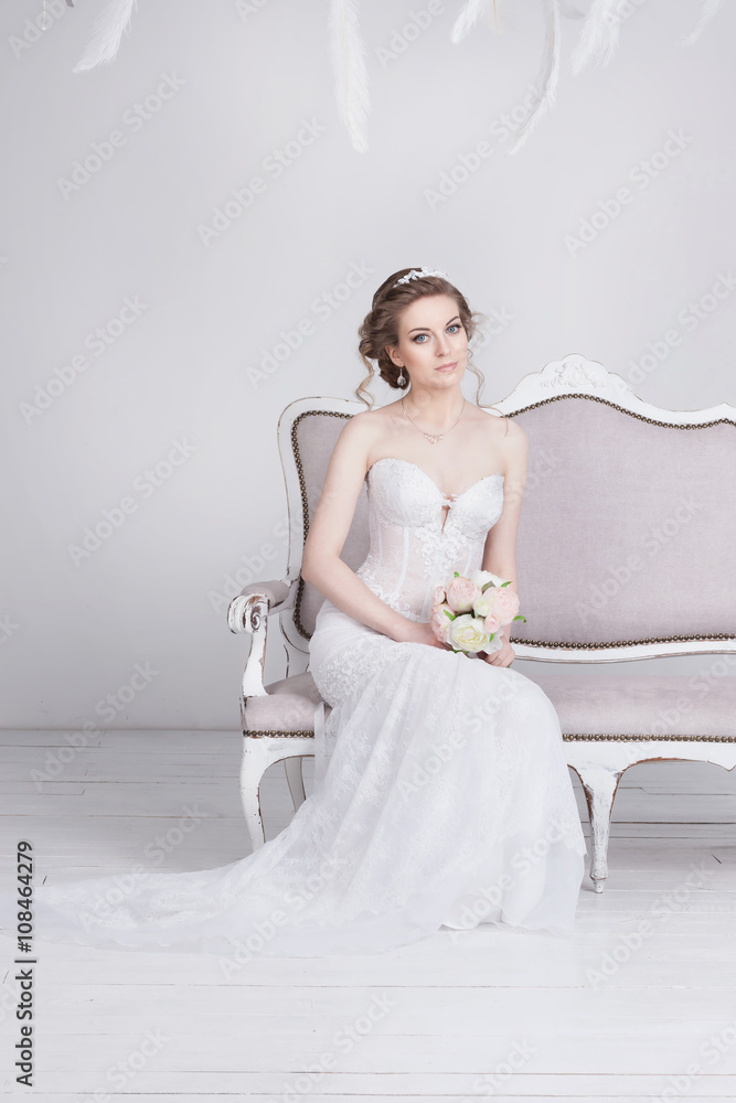 Beautiful young bride in a luxurious lace wedding dress. She sits on a  white vintage sofa.