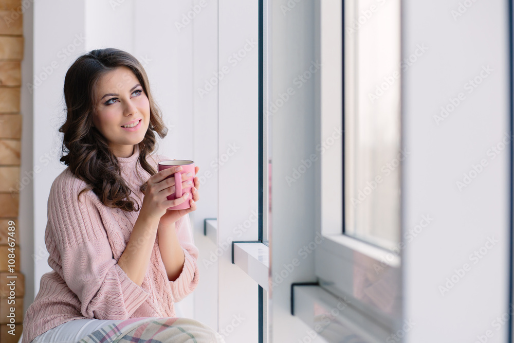 Pretty young woman in the pink sweater sitting nestled plaid and drinking coffee on the windowsill near the window Pretty young woman in the pink sweater sitting nestled plaid and drinking coffee on the windowsill near the window