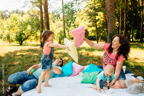 Young family with kids having fun with colored pillows outdoors. Parents with two children relax in a sunny summer garden. Mother, father, little girl and baby boy playing in park. Pillow fight © SPY_studio