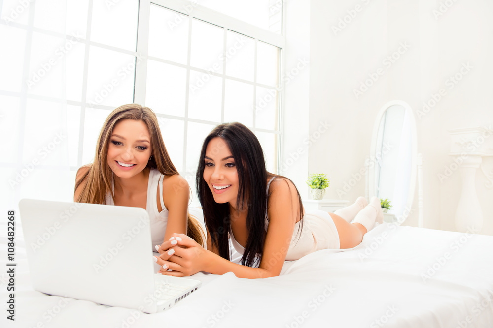 Two attractive girlfriends lying on bed with white laptop Two attractive girlfriends lying on bed with white laptop