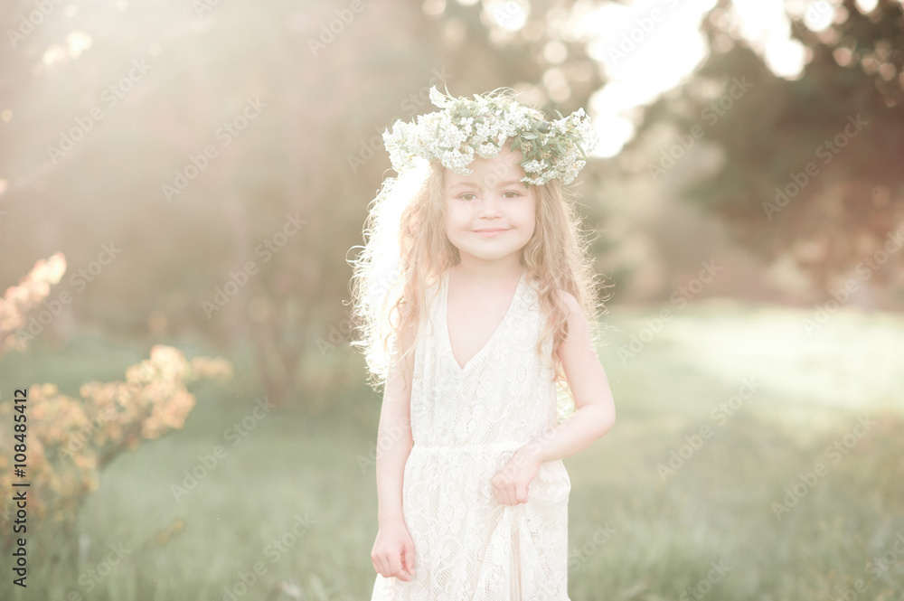 Smiling kid girl 4-5 year old wearing stylish white dress and flower wreath outdoors. Looking at camera. Sunny day. 